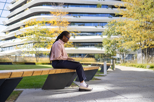 Young black female entrepreneur surfing the Internet on a computer while relaxing on a park bench in front of an office building.