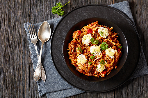 Lasagnette with beef ragu, mushrooms and ricotta cheese in black bowl on dark wooden table with spoon and fork, italian recipe, horizontal view from above, flat lay, free space