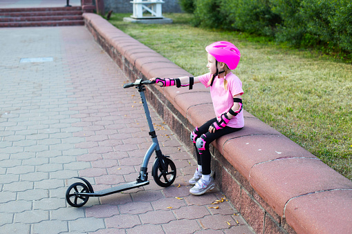 A girl in pink protective gear sits on a curb next to a scooter. The concept of safety while riding a scooter.