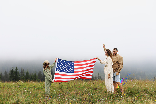 Patriotic holiday in United States of America. Happy family, parents and daughter child girl with American flag in nature on foggy mountains. The USA celebrate 4th of July