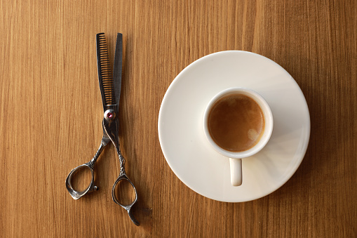 Hair cutting steel shear and white coffee cup with plate isolated on wooden background. Professional barber shop tools. Coffee break service in barbershop. Top view.