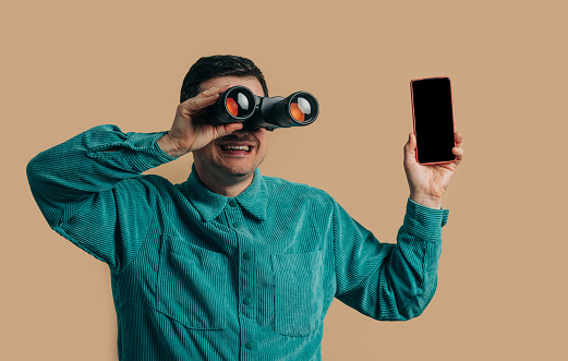 Stylish caucasian man in green shirt with smartphone and binoculars on brown background