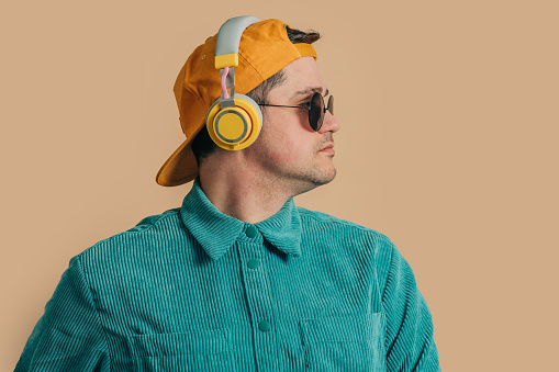 Stylish caucasian man in green shirt, headphones, sunglasses and cap on brown background