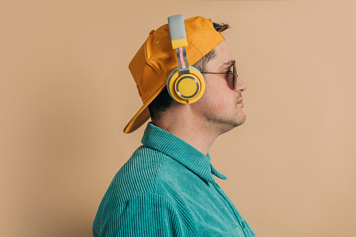 Stylish caucasian man in green shirt, headphones, sunglasses and cap on brown background