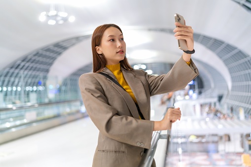 Asian woman with suitcase using smartphone at the airport terminal departure lounge boarding gate while waiting for her flight. Young woman on business trip. Young Asian Woman Digital Banking Anytime, Anywhere. Lifestyle and technology. Business on the go.