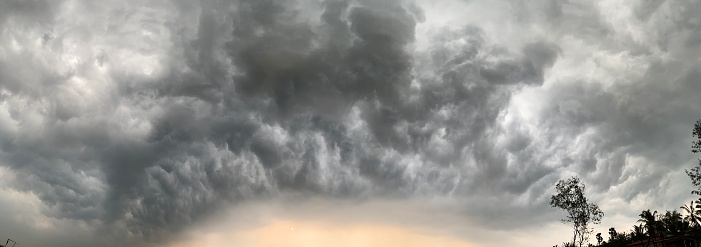 Panoramic cloudscape of dark monsoon clouds in the sky above a silhouette of trees.