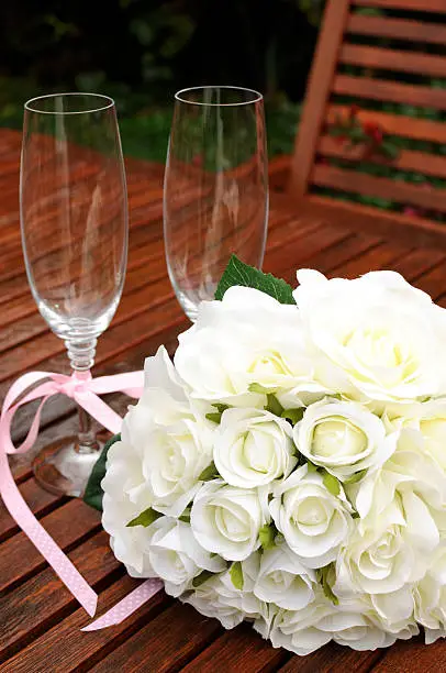 Wedding bridaal bouquet of white roses with two champagne glasses with pink polka dot ribbon on outdoor garden table setting after rain. Vertical.