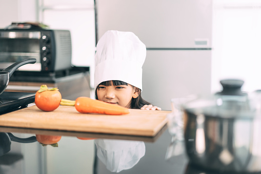 Little asian girl with chef hat and apron cooking in kitchen with recipe. Leisure lifestyles with homemade food at home. Kid growth with interest skill concept.
