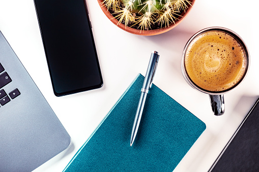 Desk, top view on a white background. Coffee, notebook and pen, phone, plant, and laptop, overhead flat lay shot