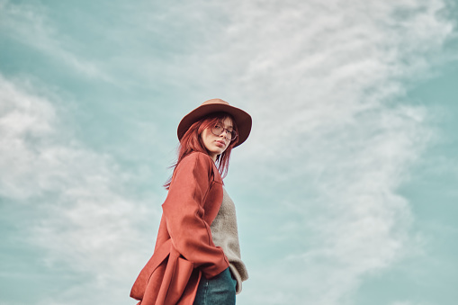 Confident teenage girl in red coat and hat standing against the sky with hands in pockets and looking at the camera over her shoulder. Low angle view.