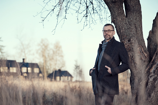 Man in a coat stands near a tree in a park in the countryside. Rest, slowing down, meditative mood.