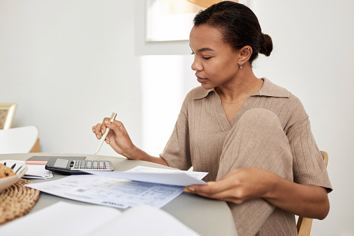 Minimal side view portrait of young Black woman doing taxes at home and using calculator, copy space