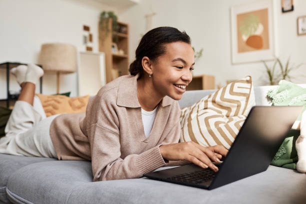Smiling Black girl using laptop lying on couch in cozy home