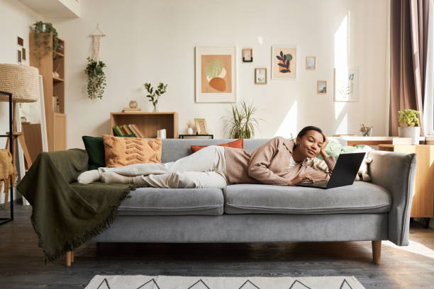 Young Black woman lying on couch in cozy home interior and using laptop