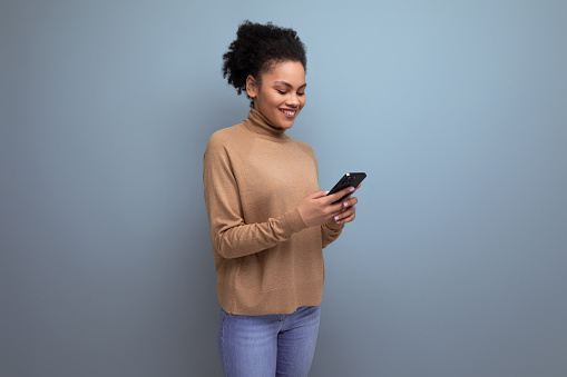 positive pretty young woman with dark skin and fluffy hair chatting online in smartphone over isolated background with copy space.