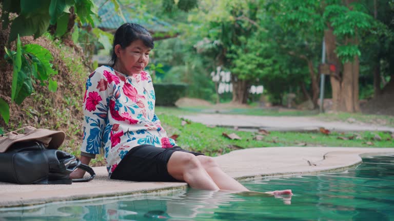 Senior Asian female tourist relaxing near swimming pool by sitting near poolside while her legs putting in the pool at a resort. Carefree senior woman spending leisure time enjoying tranquility with nature environment.