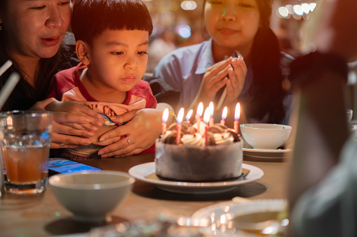 Cropped view of An Asian litte cute boy happy and his family on celebrate birthday with festive cake blowing party at home. Celebrate concept. Selective focus at boy.