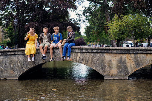 Mother and three teenage kids enjoying summer vacations in Gloucestershire, United Kingdom. 
They are sitting on a small bridge over river Windrush in the beautiful village of Bourton-on-the-Water.
Shot with Canon R5