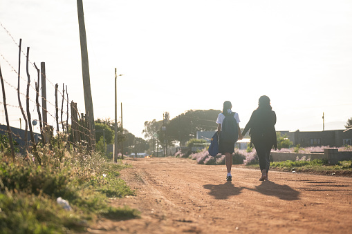 Early morning sunlight catches a mother walking with her daughter to the local school through the township. Early morning commute to school by foot.