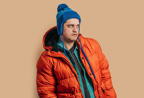 Funny caucasian guy in blue hat, orange down jacket on brown background
