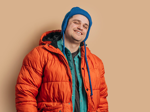 Funny caucasian guy in blue hat, orange down jacket on brown background