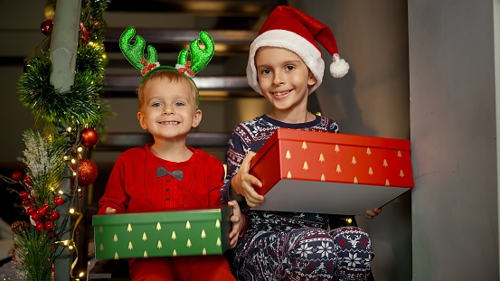 Two cheerful boys in pajamas laughing and excited after getting Christmas gifts from Santa on New Year sitting on wooden stairs at house. Family celebrations on winter holidays