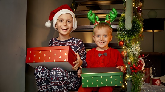 Two cheerful brothers in pajamas sitting on stairs and holding Christmas gifts and presents from Santa at morning. Family celebrations on winter holidays