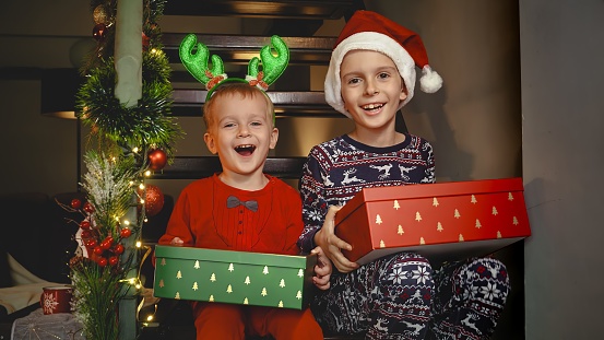 Portrait of two smiling boys in pajamas sitting on stairs with Christmas gifts and laughing in camera.