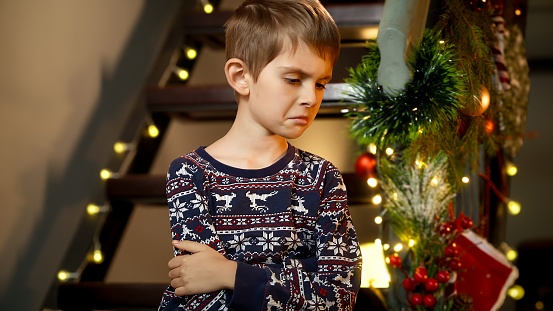 Portrait of upset unhappy boy sitting alone on wooden stairs while waiting for Santa to come on Christmas night.
