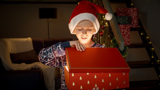 Excited little boy in Santa's hat opens magical glowing Christmas gift box with present on Christmas eve. Family celebrations on winter holidays.