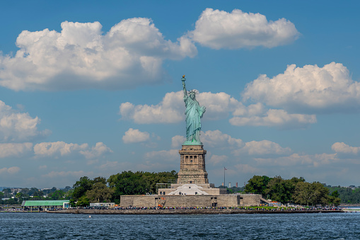Statue of Liberty, New York, USA - September 16, 2023.  A landsape panorama of The Statue of Liberty and Liberty Island full of tourists on a sunny day with white clouds and blue sky