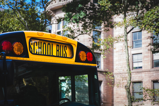 A close up of the front end of a traditional American yellow school bus outside an elementary school with large sign and safety lights in a city environment