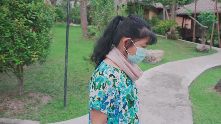 Senior Asian woman wearing facemask and casual clothing walking in a garden of a tourist resort in the morning for exercising. Female spending her leisure time relaxing in the environment surrounded by nature.