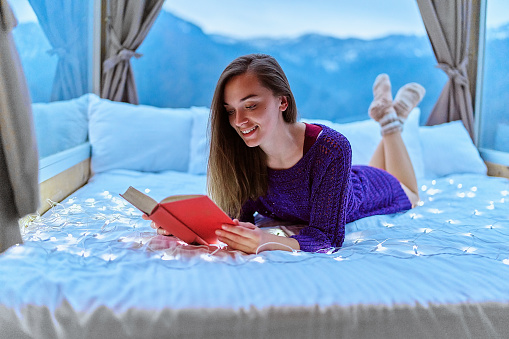 Young beautiful charming happy cute smiling romantic woman passionate about reading exciting book novel while lying on bed with mountain view