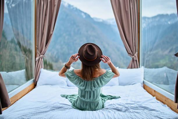 istock Young charming beautiful boho chic brunette woman traveler wearing bare shoulder emerald dress and felt brown hat sitting on bed with mountain view 1791396866