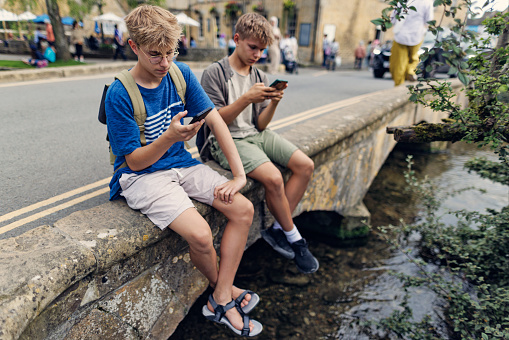 Teenage boys enjoying summer vacations in Gloucestershire, United Kingdom. \nThey are more interested in checking smartphones then the beautiful village of Bourton-on-the-Water.\nShot with Canon R5