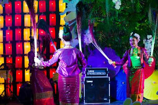 Female thai dance performance  with peacock feathers. A group of young  thai women are dancing traditional performance in traditional costumes -  they are performing classical dance in public to people who visit square and night market  in Chiang Mai. All dancers are holding peacock feathers,  Scene is at One Nimman Square. Performance is on a small stage and they are dancing in public for people who are visiting market as part of several entertainment of dancers and singers