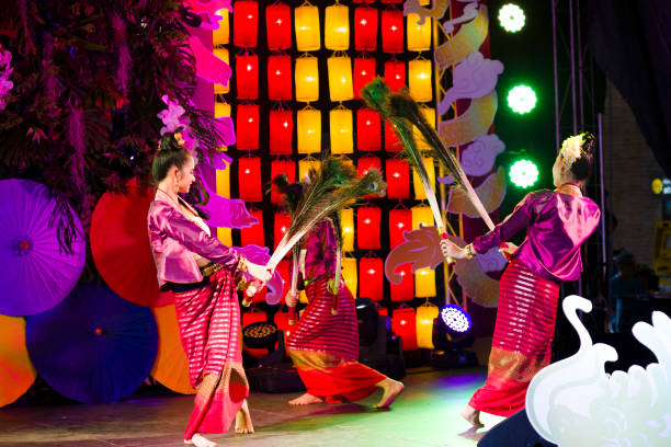 Female thai dancers are dancing Rum Thai  in a circle  with peacock feathers. Female thai dancers are dancing Rum Thai  in a circle  with peacock feathers. Young  thai women are dancing traditional performance in traditional costumes in public to people who visit square and night market  in Chiang Mai. All dancers are holding peacock feathers,  Scene is at One Nimman Square. Performance is on a small stage and they are dancing in public for people who are visiting market as part of several entertainment of dancers and singers true thailand classic stock pictures, royalty-free photos & images