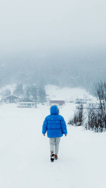 donna in cappotto blu che ammira la fiaba invernale che cammina nel villaggio scandinavo durante una forte nevicata - rear view winter blizzard nordic countries foto e immagini stock