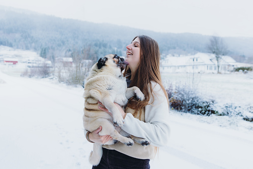 Portrait of a smiling female with long hair embracing with a pug on the village street with view of the snowcapped mountains in Scandinavia