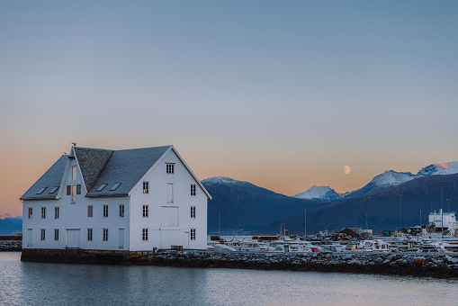 Dramatic view of the Moon in the twilight sky above the city harbour and snowy mountain peaks of Norway, Scandinavia