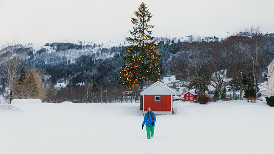 Front view of happy female in colourful warm clothing contemplating the Christmas time walking to the old wooden hut with view of the green pine tree in Christmas lights and snowcapped mountain peaks in Norway