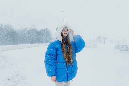 Portrait of female wearing warm winter jacket walking in the town during strong winter blizzard by the road with cars driving through storm contemplating Christmas time outdoors