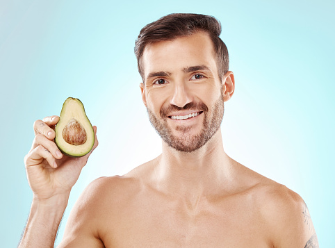 Man, skincare and studio portrait with avocado for health, nutrition and cosmetic wellness by blue background. Happy young model, fruit and healthcare for natural detox, facial skin glow or aesthetic