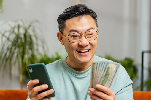 Rich happy Asian man planning budget use smartphone calculator app count money cash calculate domestic bills at home. Mature guy satisfied of income earnings win saves money for planned vacation gifts