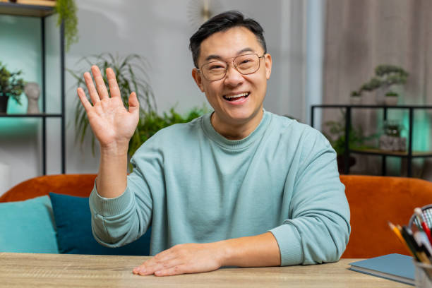 istock Asian man smiling friendly at camera, waving hands gesturing hello, hi, greeting at home on sofa 1791525779