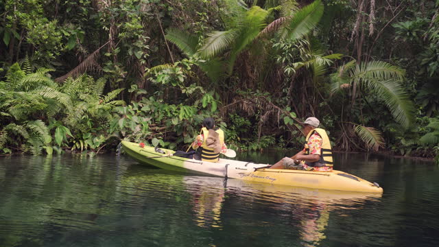 Senior adult Asian tourist couple wearing casual clothing and life jacket traveling together by kayaking into a mangrove forest and taking photos for capturing a beautiful moment and beauty of nature.