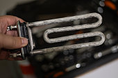 A hand holds a heating element with limescale deposits in front of a washing machine. Damage to the washing machine due to the accumulation of limescale on the heating element.