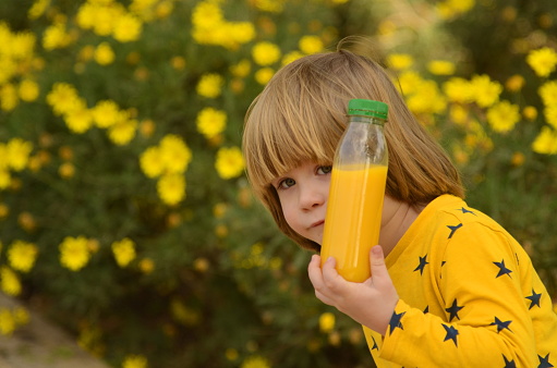 A cute baby in a yellow sweater is holding a plastic bottle with fresh orange juice. The boy walks in nature, drinks freshly squeezed juice. Concept: healthy natural organic juice