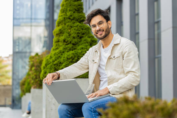 Indian man freelancer start working opens laptop sends messages reading email outdoors on bench